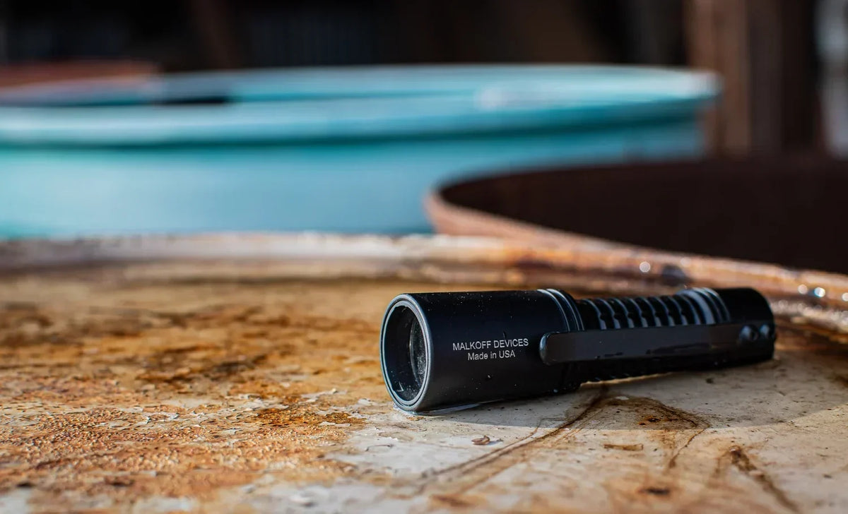 Black Malkoff Devices LED flashlight resting on a rusty surface outdoors